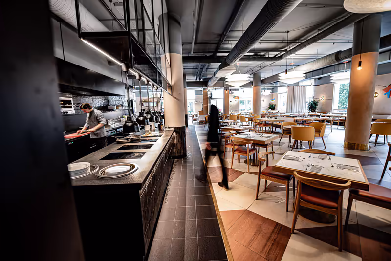 Modernes Restaurant-Interior mit offener Küche, Koch am Herd, Servicepersonal und gedeckten Tischen, helles Loftdesign