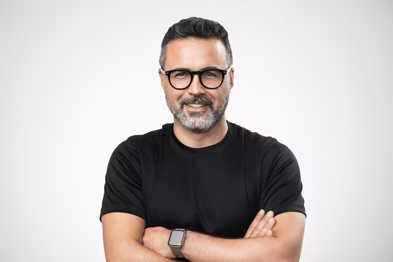 Mann mit Brille und schwarzem T-Shirt, verschränkte Arme, selbstbewusstes Lächeln, Portrait vor neutralem Hintergrund