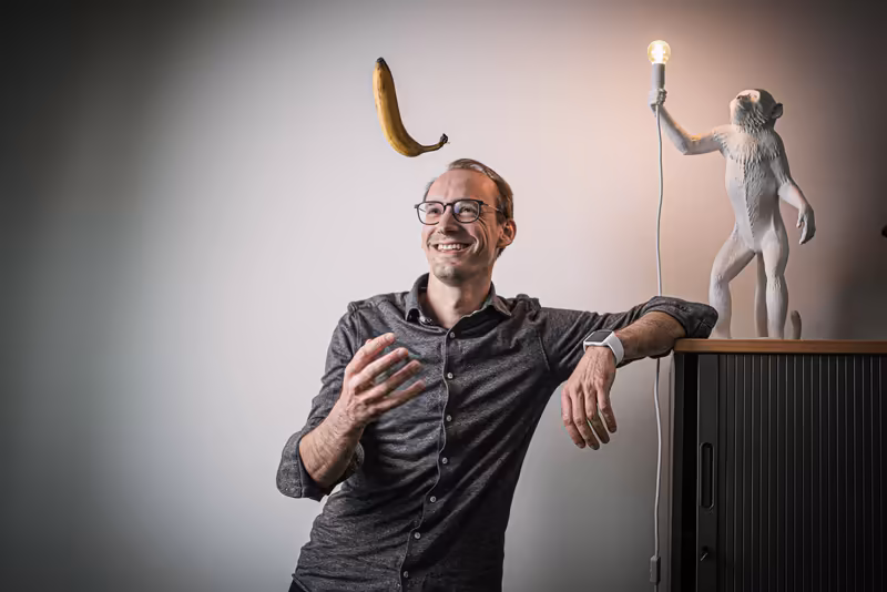 Mann mit Brille jongliert Banane, neben ihm Affenskulptur mit Lampe, humorvolle Business-Porträtaufnahme