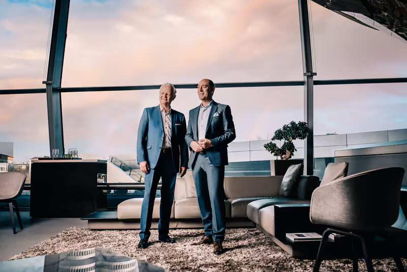 Zwei Geschäftsmänner in Anzügen stehen in moderner Penthouse-Lounge mit Panoramafenstern, Skyline-Ausblick und designtem Interieur