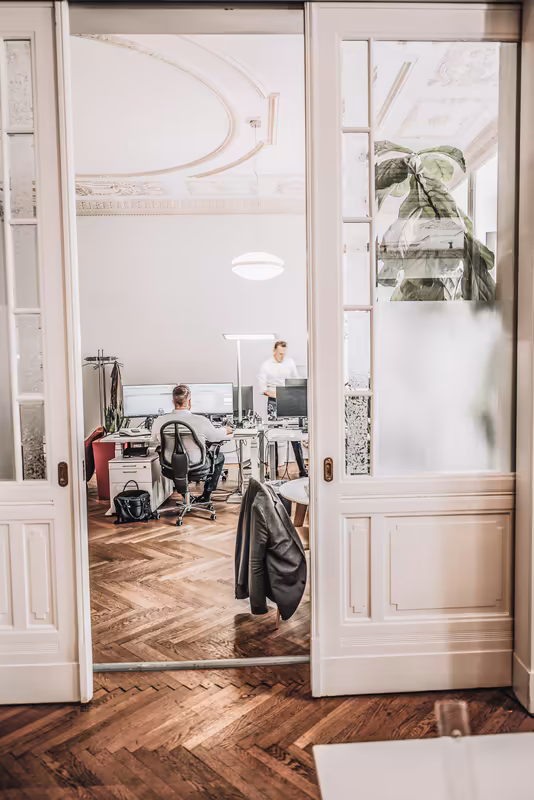 Modernes Büro mit zwei Personen am Schreibtisch, Blick durch offene weiße Türen, helles Licht, Pflanze, Parkett