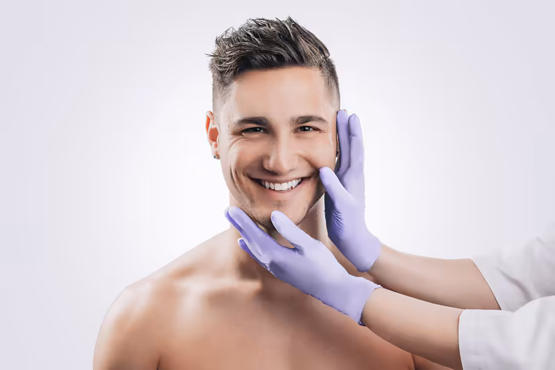 Mann bei dermatologischer Untersuchung, lächelnd, mit violetten Handschuhen des Arztes im Gesicht