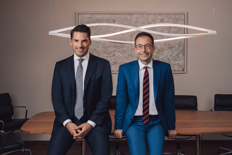 Zwei Geschäftsmänner in Anzügen sitzen an Konferenztisch in modernem Büro, selbstbewusste Businessfotografie