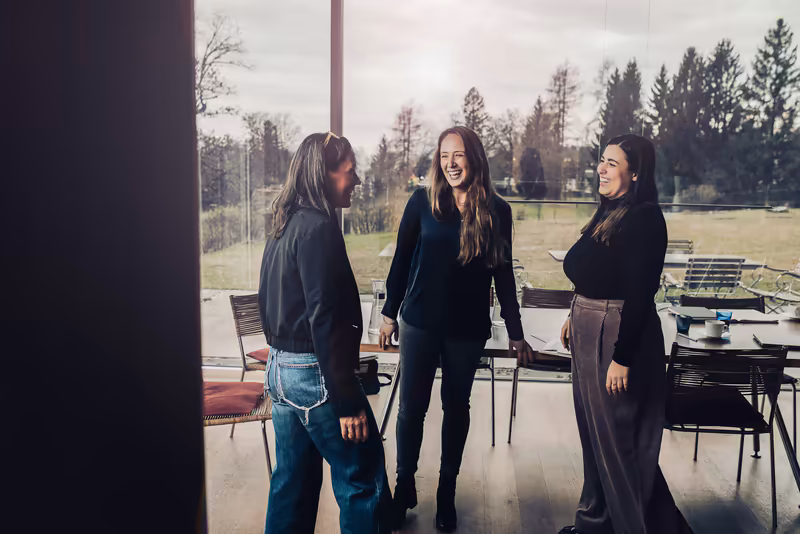 Drei Geschäftsfrauen in schwarzer Kleidung unterhalten sich lachend in moderner Bürolounge mit Panoramafenstern und Waldblick