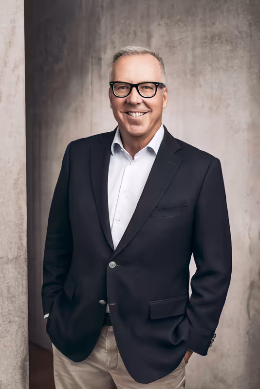 Mann im schwarzen Blazer und weißem Hemd mit Brille, lächelnd vor grauer Betonwand, Business-Porträt
