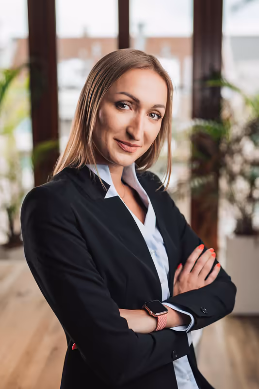 Geschaeftsfrau in schwarzem Blazer mit verschraenkten Armen, Portrait in modernem Buero mit Glasfassade und Pflanzen