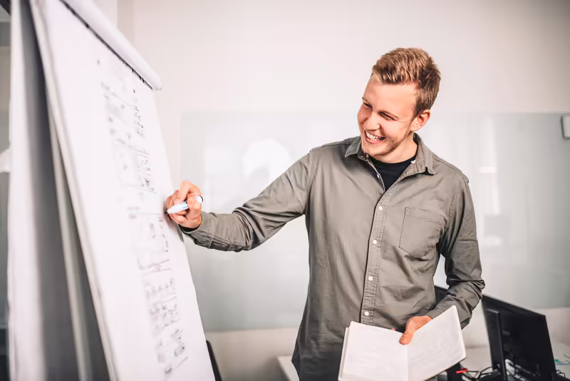 Mann in grauem Hemd präsentiert lächelnd Skizzen auf Whiteboard, Business-Umgebung, Brainstorming-Szenario