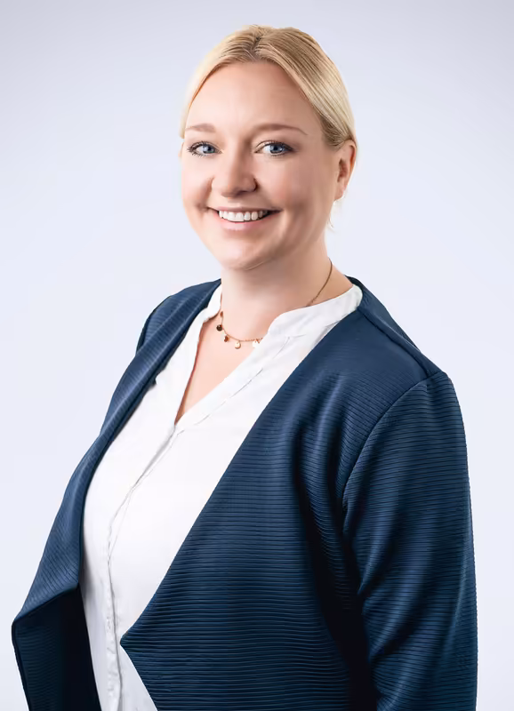 Professionelles Porträt einer Frau in dunklem Businessblazer über weißer Bluse, lächelnd vor grauem Hintergrund