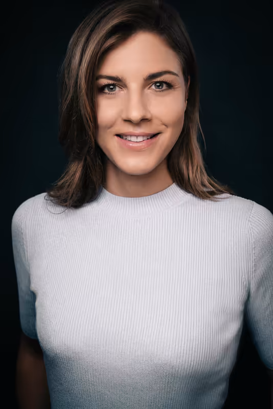 Porträt einer Frau mit braunen Haaren, weißem Strickpullover, lächelnd vor dunklem Hintergrund, Business-Headshot