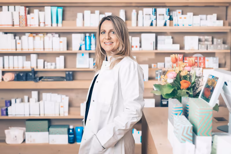 Frau in weißem Blazer an Schreibtisch, Regale mit Produkten im Hintergrund, professionelles Porträt in Beauty- oder Wellness-Umgebung