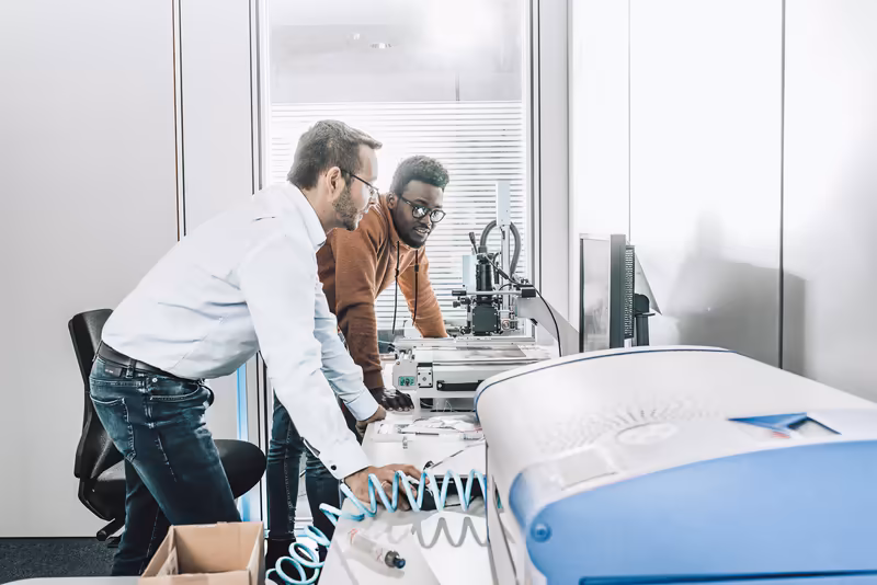 Zwei Männer arbeiten konzentriert an 3D-Drucker und technischen Geräten in modernem Büro mit Glasfenster