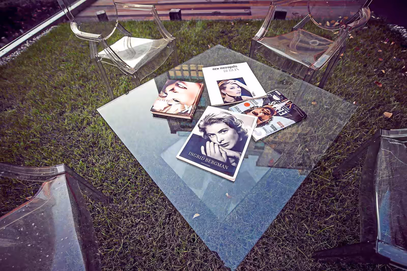 Overhead-Aufnahme: Glasplattform mit Printmaterialien und Porträts auf Grünfläche, zwei transparente Stühle, professionelle Businesspräsentation