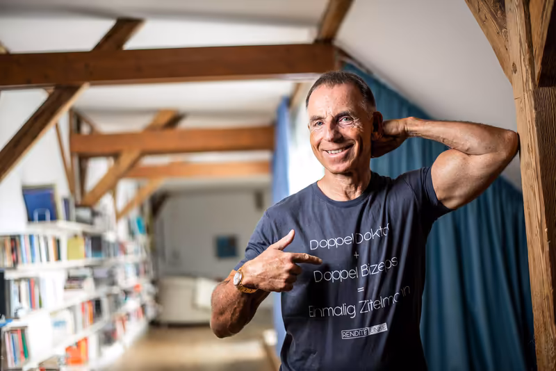 Mann in dunklem T-Shirt lächelt in moderner Loft-Büroumgebung mit Holzbalken und Bücherregal, lässige Pose