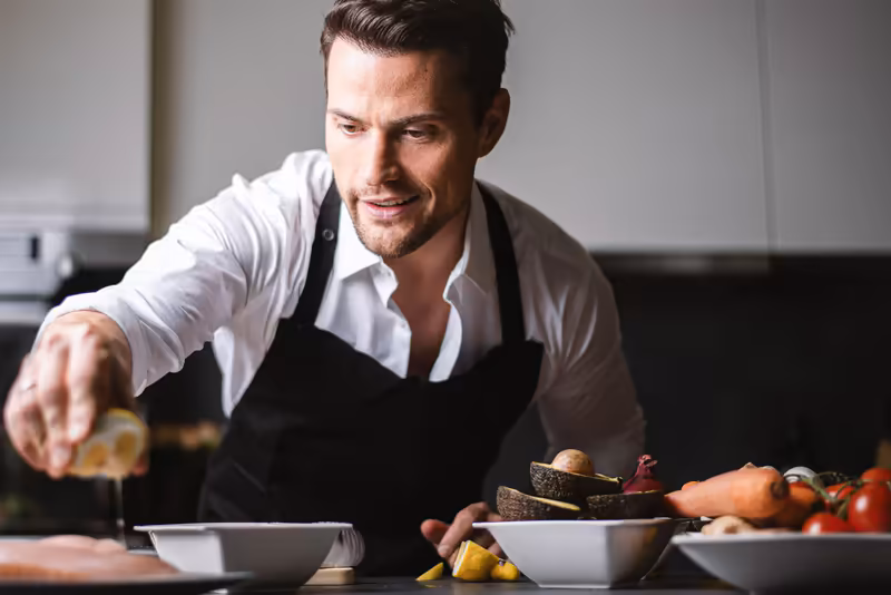 Mann in weißem Hemd und schwarzer Schürze bereitet Speisen in moderner Küche zu, professionelle Gastronomie-Portrait