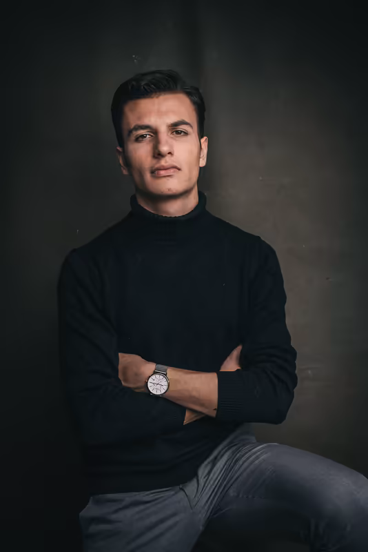 Mann in schwarzem Rollkragenpullover sitzt mit verschränkten Armen, trägt Armbanduhr, ernster Blick, dunkler Hintergrund