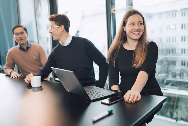 Drei Geschäftsleute an Schreibtisch mit Laptop in modernem Büro mit Panoramafenstern, freundliche Arbeitsatmosphäre