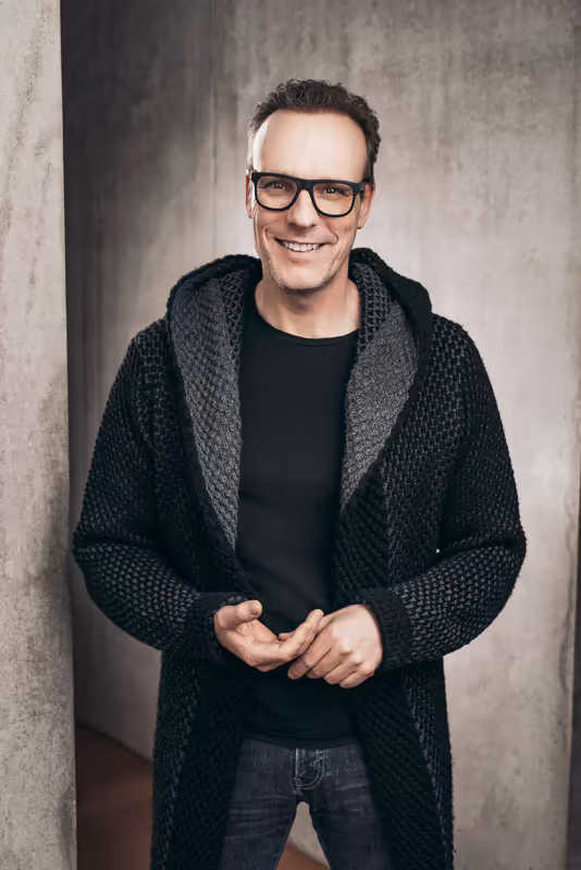 Mann mit Brille in grünem Strickmantel vor Betonwand, lächelnd, Business-Porträt
