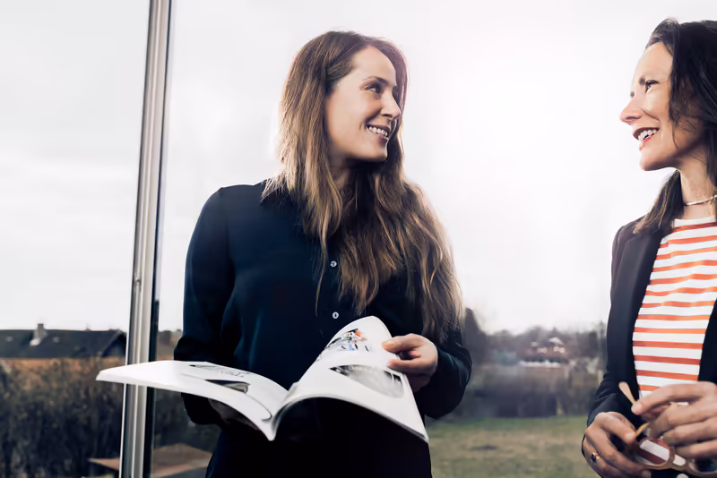 Zwei Frauen im Business-Outfit lachen zusammen, eine hält eine Zeitschrift, Fenster mit Landschaftsblick