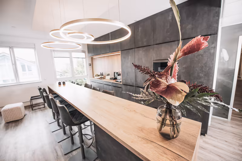 Modernes Büro mit Konferenztisch, schwarzer Küchenzeile, Designleuchten und Blumenarrangement auf Holztisch