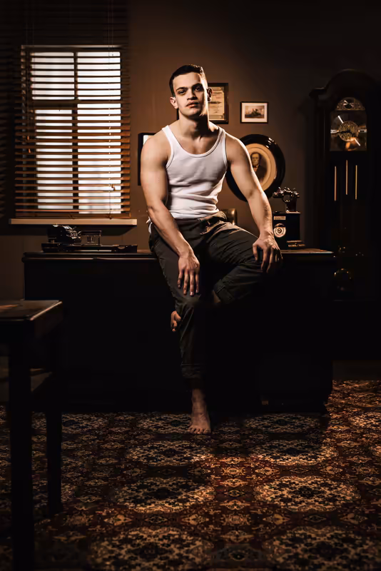 Mann sitzt auf Schreibtisch in warmem Vintage-Interieur, weißes Unterhemd, dunkle Hose, nachdenkliche Pose, Fenster mit Jalousien im Hintergrund