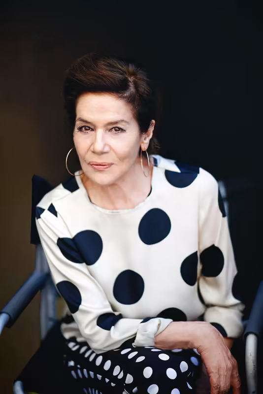 Frau mit dunklem Haar und Creolen sitzt auf Stuhl, trägt cremefarbenes Oberteil mit dunklen Punkten, ruhiger Blick, dunkler Hintergrund