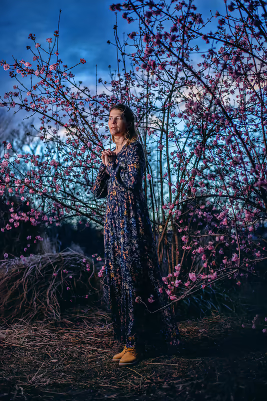 Frau steht zwischen blühenden Sträuchern mit rosa Blüten in der Abenddämmerung, nachdenkliche Atmosphäre