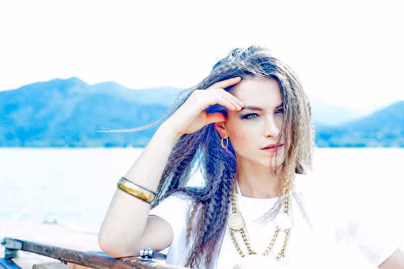 Junge Frau mit geflochtenen Haaren am See, Hand ins Haar, bergige Landschaft im Hintergrund, nachdenklicher Blick