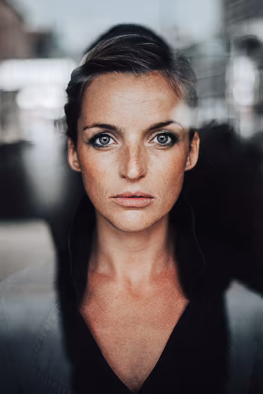 Porträt einer Frau mit direktem Blick, schwarzem Haar und ernster Mimik vor unscharfem urbanem Hintergrund