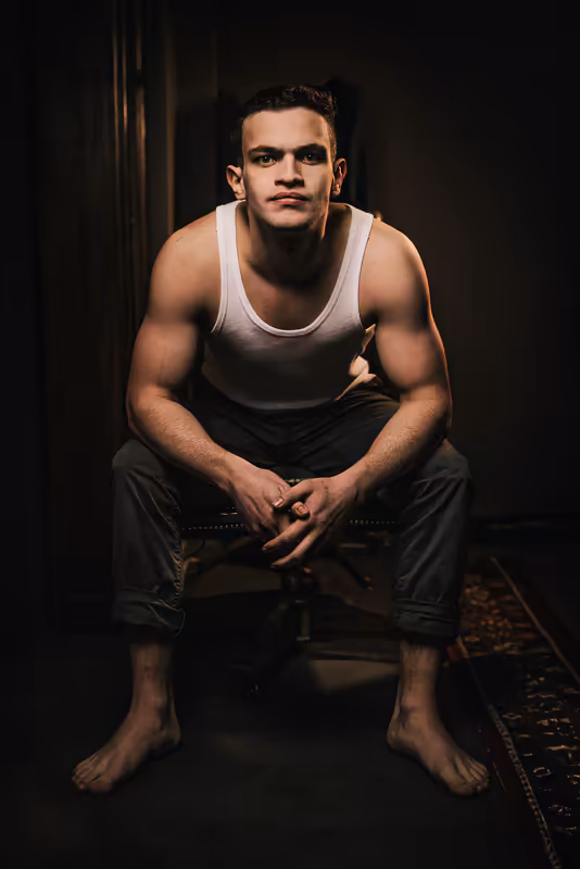 Porträt eines muskulösen Mannes im weißen Tanktop, sitzend vor dunklem Hintergrund, ernster nachdenklicher Blick