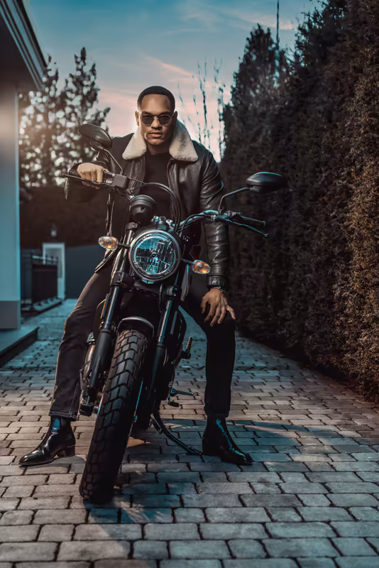 Mann mit Sonnenbrille sitzt auf Motorrad in enger Gasse, schwarze Lederjacke, Sonnenuntergang, urbane Stimmung