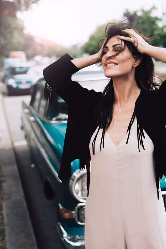 Frau lehnt an Vintage-Auto, blickt zur Seite, Hand im Haar, entspannte Atmosphäre auf Straße