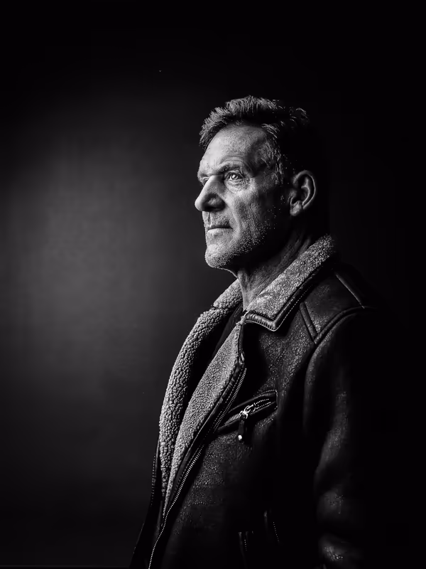 Porträt eines Mannes im Profil, Schwarzweiß-Aufnahme, ernste Mimik, Lederjacke mit Wollkragen, dramatische Beleuchtung vor dunklem Hintergrund