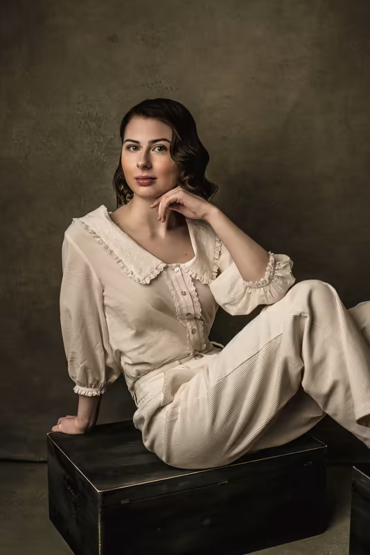 Frau in cremefarbener Vintage-Bluse sitzt auf schwarzem Koffer, nachdenklicher Blick, neutraler Hintergrund