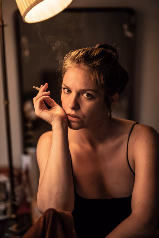 Frau mit Zigarette unter Studiolampe, nachdenklicher Blick, warmes Licht, dramatische Porträtfotografie