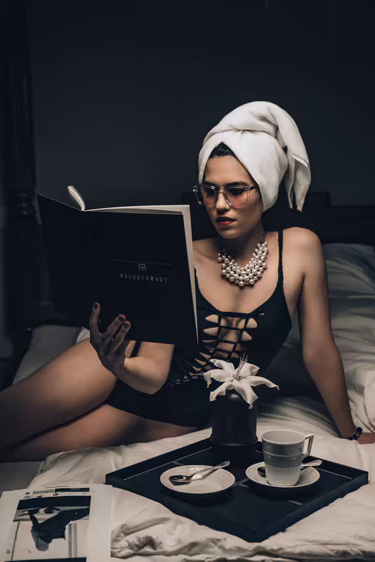 Frau mit Turban und Brille liest Buch im Bett, Perlenkette, schwarzes Top, Tablett mit Tasse und Blume, dunkle Studioatmosphäre